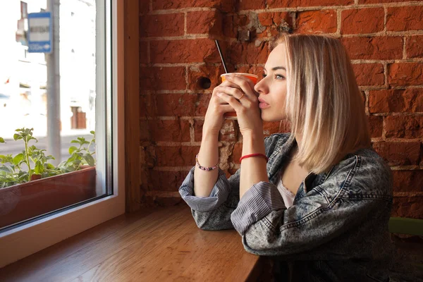 Young blonde woman holding orange paper cup with cocktail straw sitting near window against red brick wall at the cafe dreamily looking out the window