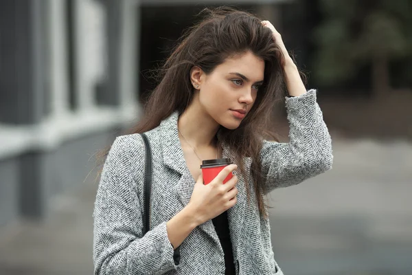 Lifestyle fashion portrait of beautiful young brunette woman in grey coat with coffee cup posing on street cloudy day