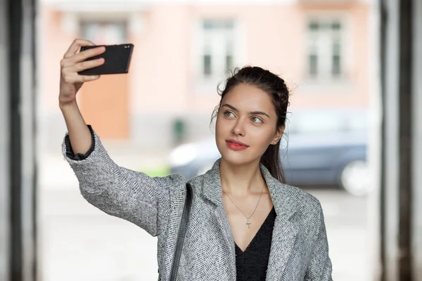 Beautiful young brunette woman in grey coat taking a selfie with her smartphone on city street cloudy day