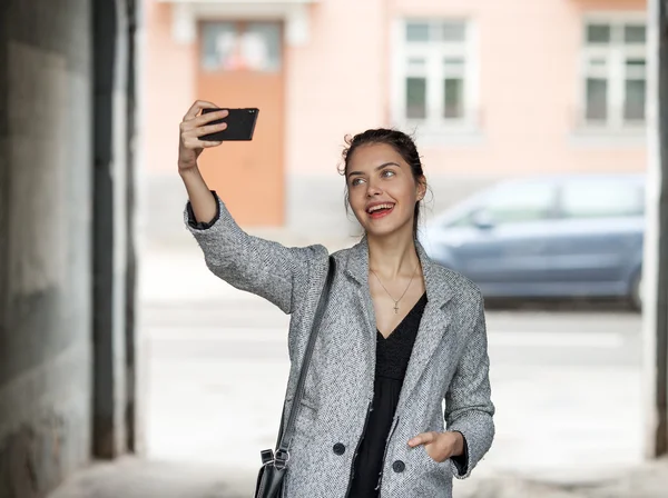 Beautiful young brunette woman in grey coat taking a selfie with her smartphone on city street cloudy day