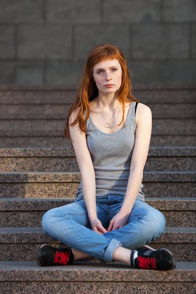 Young redhead caucasian woman in grey shirt blue jeans black sneakers sitting on stairs outdoor portrait
