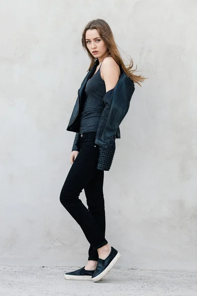 Happy young beautiful woman in black leather jacket black jeans slip-on posing for model tests against textured wall