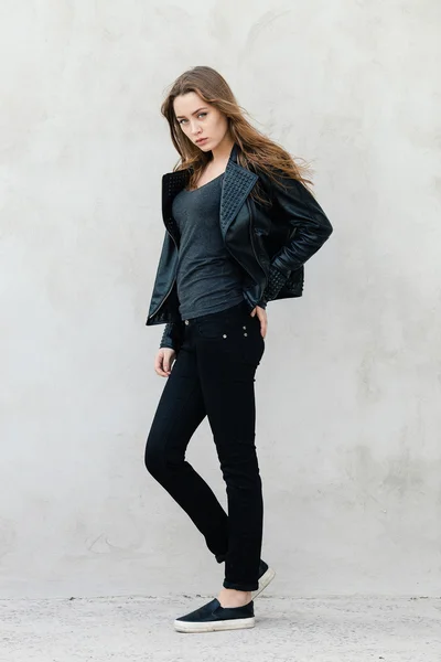 Happy young beautiful woman in black leather jacket black jeans slip-on posing for model tests against textured wall