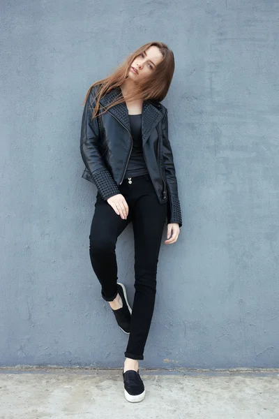 Happy young beautiful woman in black leather jacket black jeans slip-on posing for model tests against textured wall