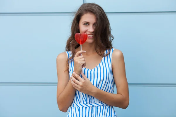 Outdoor smiling portrait of happy young beautiful brunette woman in striped summer dress posing with red heart lollipop against metal geometric background