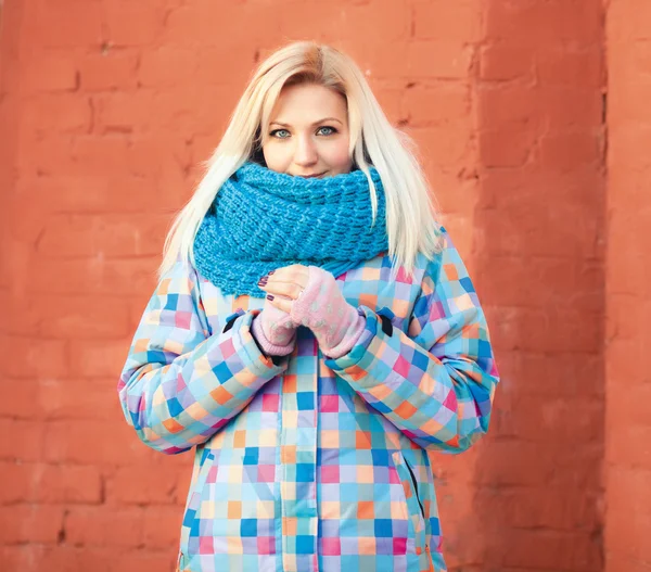 Naughty smiling beautiful blonde woman in bright jacket and scarf against a brick wall winter outdoors