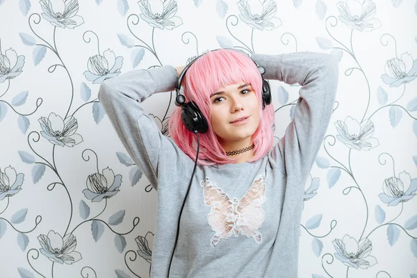 Young hipster woman in pink wig and dj headphones enjoying music against wall with vintage wallpapers pattern