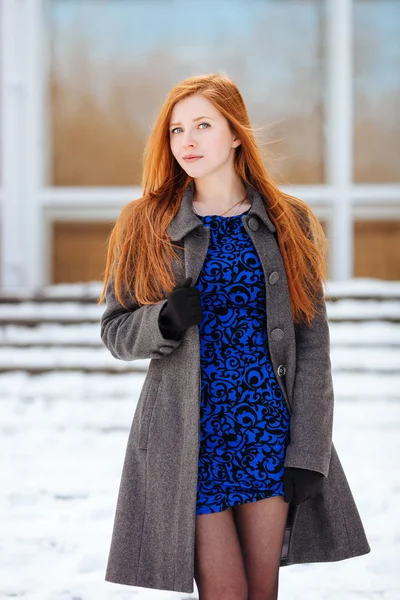 Portrait of young adorable redhead woman in blue dress and grey coat at winter outdoors