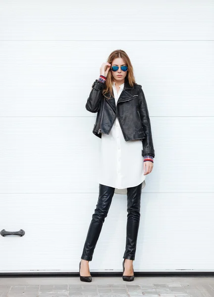 Young beautiful fashionable woman in leather jacket and white blouse posing outdoors against garage door
