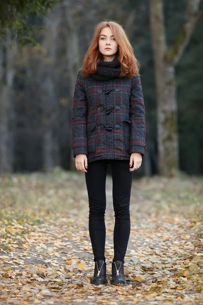 Full length outdoors portrait of young beautiful redhead woman in scarf, jacket, black jeans and boots standing on a forest path