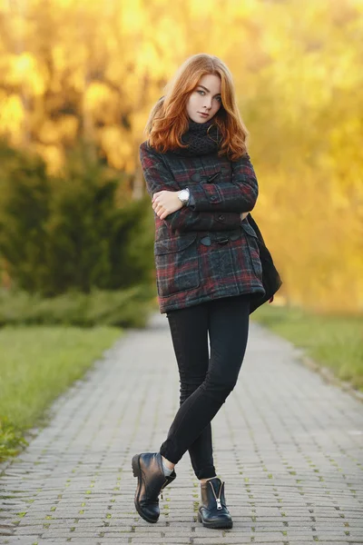 Full length portrait young trendy redhead woman in scarf and plaid jacket on park path with autumn foliage background cold season outdoors