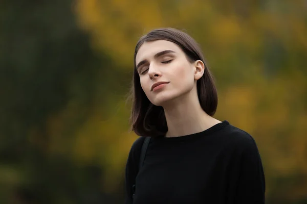 Closeup portrait of young dreamy elegant brunette hipster woman in black blouse posing with closed eyes against blurred foliage background