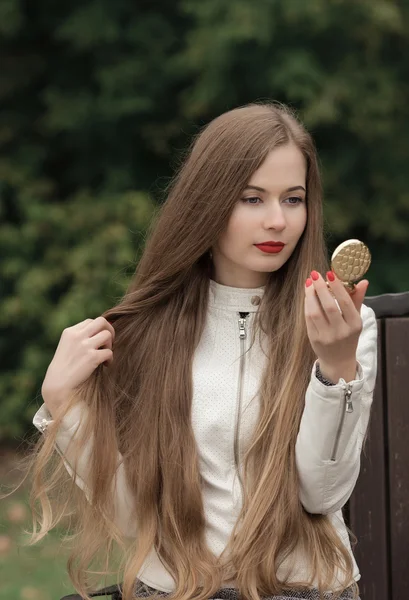 Young beautiful blonde woman with extra long straight hair bright red lipstick holding puff-box looking into mirror and corrects hairstyle posing on park bench