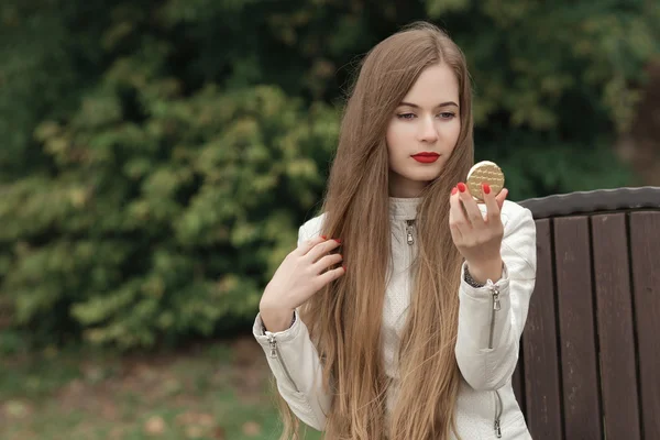 Young beautiful blonde woman with extra long straight hair bright red lipstick holding puff-box looking into mirror and corrects hairstyle posing on park bench