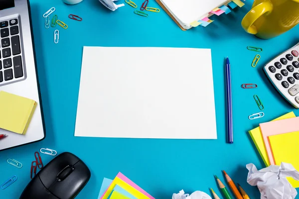 Office table desk with set of colorful supplies, white blank note pad, cup, pen, pc, crumpled paper, flower on blue background. Top view and copy space for text