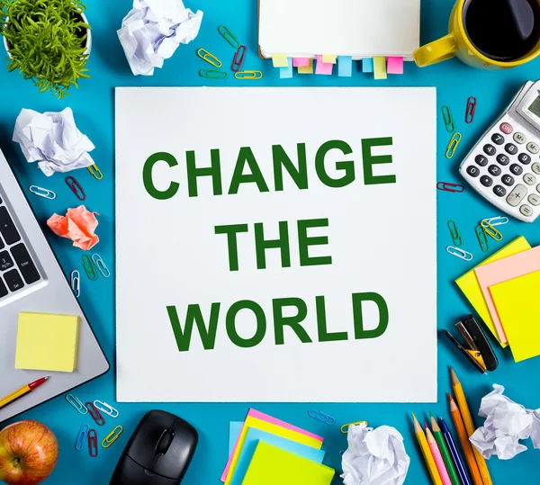 Change the world. Office table desk with supplies, white blank note pad, cup, pen, pc, crumpled paper, flower on blue background. Top view