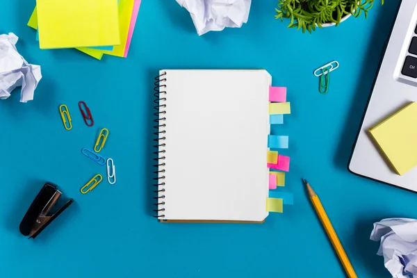 Office table desk with set of colorful supplies, white blank note pad, cup, pen, pc, crumpled paper, flower on blue background. Top view and copy space for text