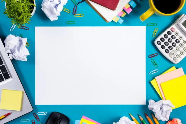 Office table desk with set of colorful supplies, white blank note pad, cup, pen, pc, crumpled paper, flower on blue background. Top view and copy space for text