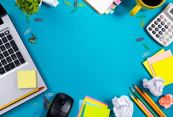 Office table desk with set of colorful supplies, white blank note pad, cup, pen, pc, crumpled paper, flower on blue background. Top view and copy space for text