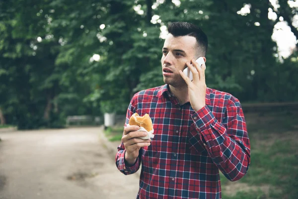 Handsome young man eating sandwich autdoor. He is holding a phone