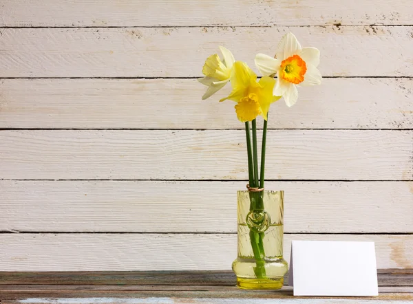 Three spring flower yellow and white daffodils with Golden wedding rings in glass vase with greeting blank card on a white wooden background