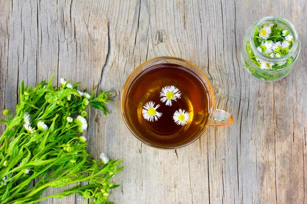 Glass cup with tea of chamomile, a bunch of chamomile flowers pharmacy and a glass jar with a camomile on old wooden table in the cracks close up, top view