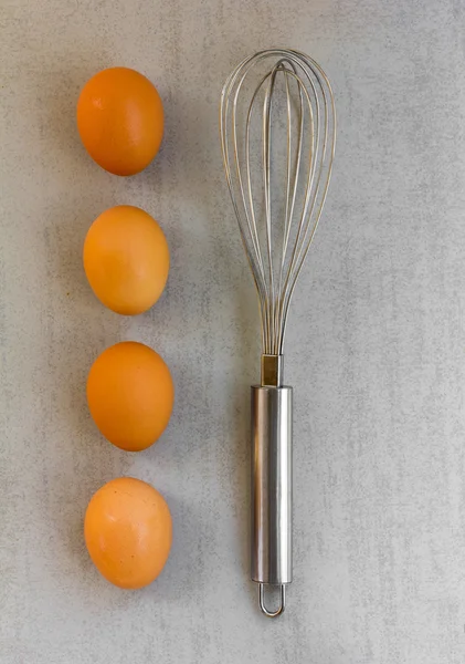 Steel whisk for whipping and mixing and raw brown eggs lined up in a row vertically on a gray background
