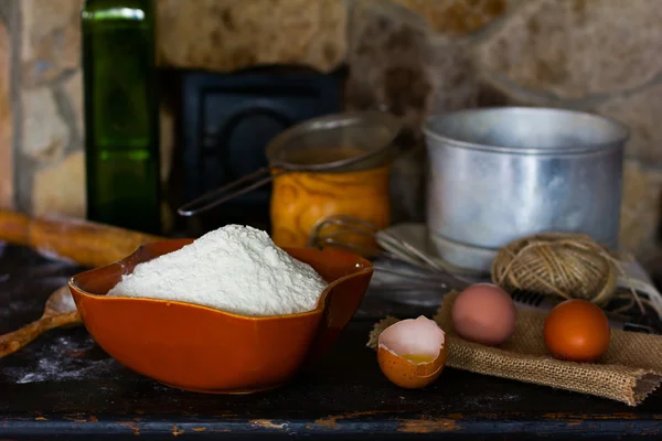 White wheat flour in ceramic ware, broken egg with the yolk, whole eggs and cooking utensils for cooking dough on a background of a stone oven. Rustic style. Side view close-up. Selective focus.