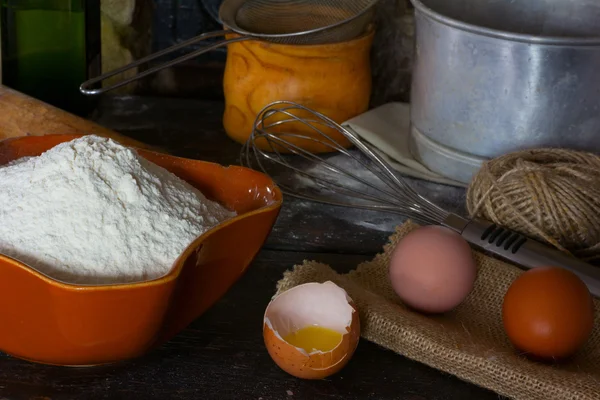 White wheat flour in ceramic ware, broken egg with the yolk, whole eggs and cooking utensils for cooking test. Rustic style. Side view close-up.