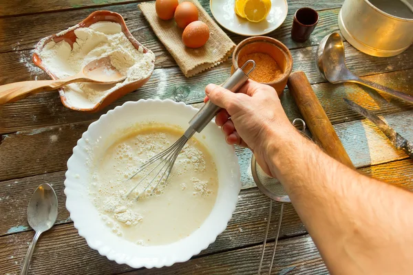 The man knead the dough with a whisk. Wheat flour, batter, eggs, a lemon and kitchen utensils on wooden table. Preparation of the dough in a rustic kitchen.