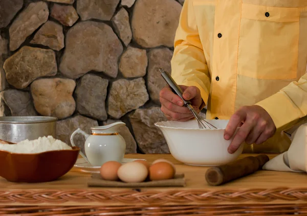 Bowl with flour, eggs, a sieve and a mortar, and a male cook in a yellow garment whisk the whisk the batter. Concept of rustic kitchen, selective focus. Copy space. Free space for text