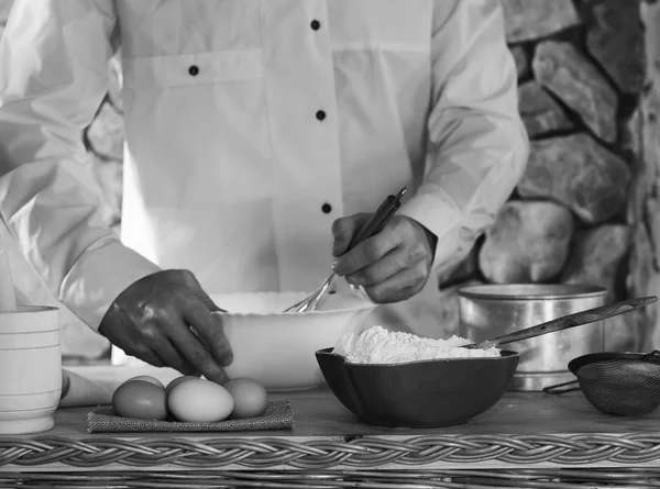 In the foreground a bowl with flour, eggs, sieve and mortar in the background a man dressed in white chef whips whisk the batter. rustic kitchen concept, selective focus. black and white photo