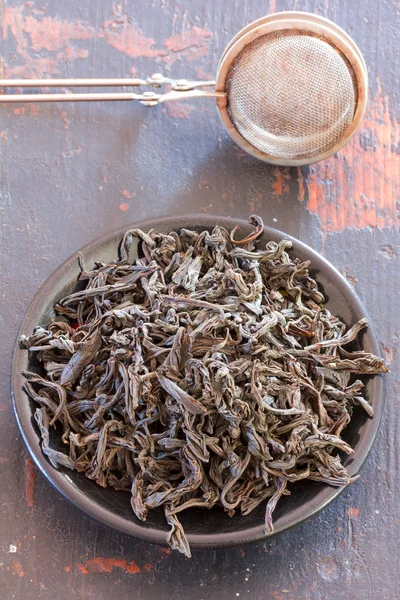 Steel strainer dried black tea leaves in a round black saucer on old wooden table close-up, top view
