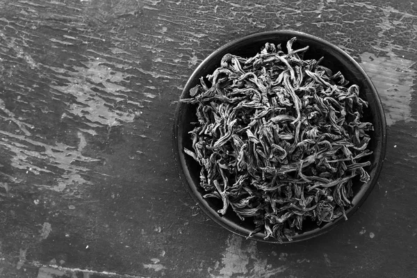 Dried black tea leaves in round black saucer on old wooden background. Copy space. Free space for text, Close-up, top view. black and white photo