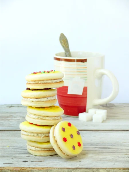 Sandwich biscuits with yellow icing sprinkled with sugar stars and cup of tea on light wooden background