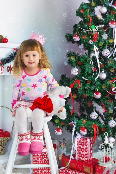 Happy little girl sitting on a chair next to the Christmas tree