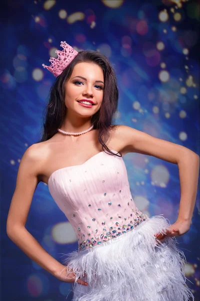 Beautiful cheerful brunette girl in a pink dress and pink crown on his head. mini with sequins and feathers. gray background.