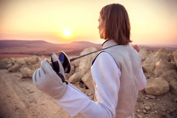 Fencer man holding his sword on the shoulders on a rocky background and looking forward to the sun goes down