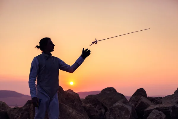 Fencer man throwing up his fencing sword on a  background of sunset sky and rocks