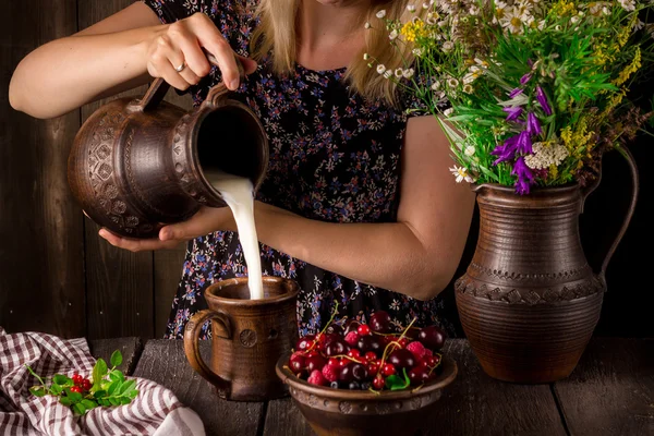The girl pouring milk from a jug into a cup and a bowl with berries on a wooden table. Jug with flowers.