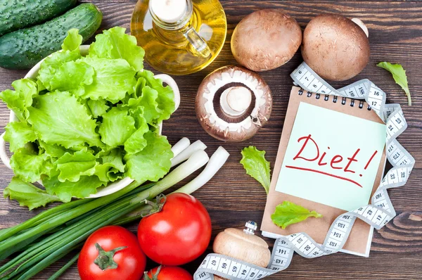 Fresh organic vegetables. Lettuce, cucumber, tomatoes, green onions, mushrooms champignons, olive oil, note Diet and measuring tape on wooden background. Healthy food and healthy life concept.
