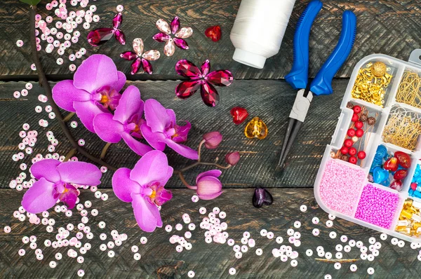 Blooming orchid flowers and set of needlework. Box with beads, spool of thread, plier and glass hearts to create handmade jewelry on old wooden background. Handmade accessories
