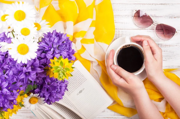 Woman's hands keep cup of coffee. Book, bouquet of flowers, female accessories - sunglasses and neckerchief on wooden table. Coffee break