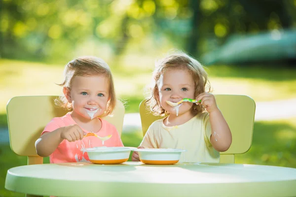 Two little girls sitting at a table and eating together against green lawn