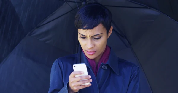 Black woman with umbrella standing in rain using mobile phone