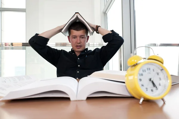 Closeup portrait of young man surrounded by tons of books, alarm clock, stressed from project deadline, study, exams. Negative emotion facial expression feelings, body language