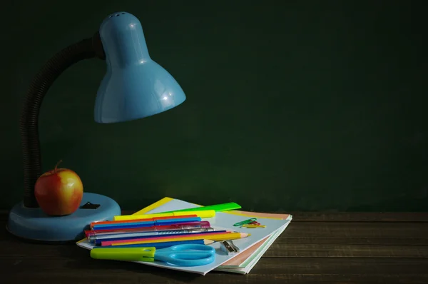 School supplies and blue desk lamp on a wooden surface against a blackboard.