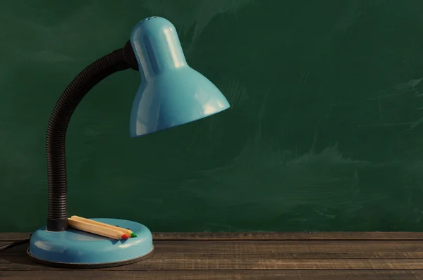 Desk lamp and colored pencils on a wooden surface against a blackboard.