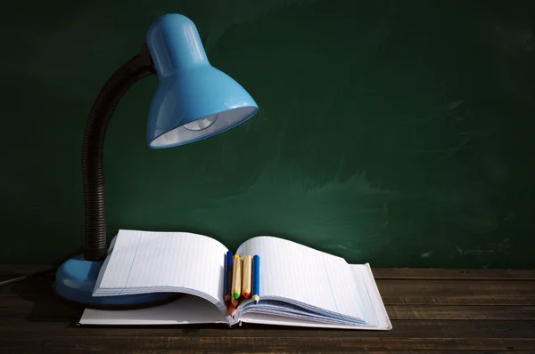 Desk blue lamp, open notebook and colored pencils against a school green board.