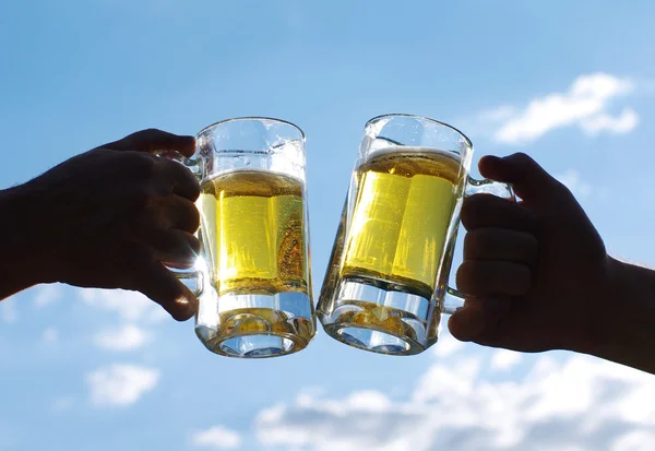 Two glasses of light beer hold male hands against the blue sky. Friends toasting with wheat beer